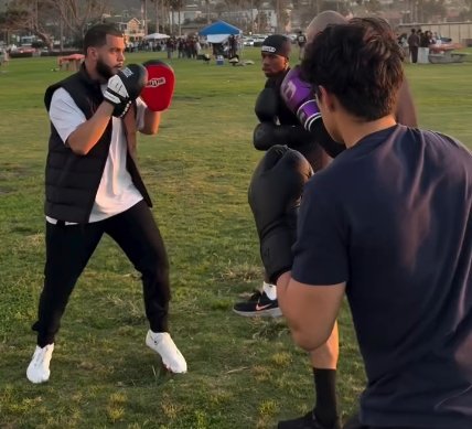 A man wearing boxing gloves and a black vest practices pad work drills outdoors
